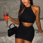 Woman in a black halter crop top and matching mini skirt, holding a red cocktail in a flute beside a black clutch on a marble bar.